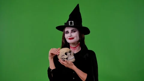 A vampire witch in a black cloak and hat poses with a skull on a green chroma Stock Footage 316588075