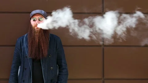 Vape man. Bearded man in a cap and sunglasses smokes an electronic cigarette. Stock Footage 107984316