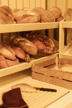 Various types of artisan bread rest on white wooden shelves in a warm baker.. Stock Photos