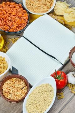 Various uncooked pasta with notebook and vegetables on a wooden table Stock Photos