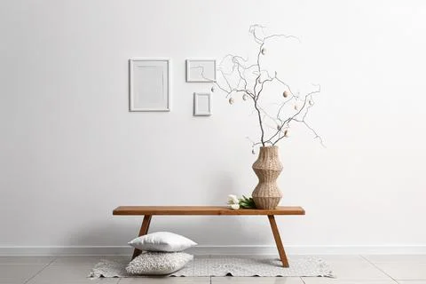 Vase with tree branches, Easter eggs and tulips on bench near light wall in r Stock Photos