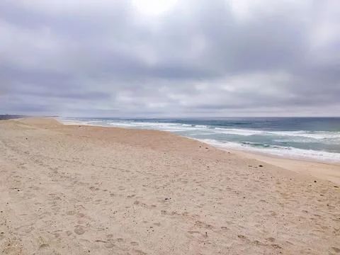 A vast, empty sandy beach stretches under a dramatic, overcast sky with rolli Stock Photos