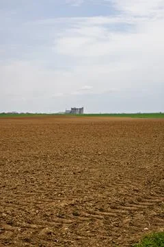 Vast Plowed Field With Distant Grain Silos Under a Wide Cloudy Sky in Rural.. Stock Photos
