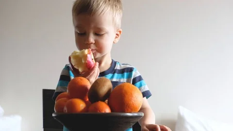 Vegetarian child eats and chews apple, plate with fresh fruits in front of him Stockbeeldmateriaal 126998267