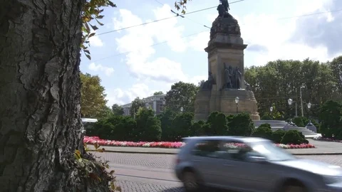 Vehicles moving around the traffic circle with sculpture, hedges and flowers Stock Footage 73702404