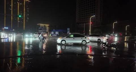 Vehicles navigate a reflective, rain-soaked city intersection at night. Stock Footage 336199789