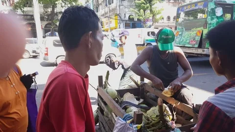 Vendor with Myanmar cap cuts coconut, coconut cart, traffic on road, costumers Stock Footage