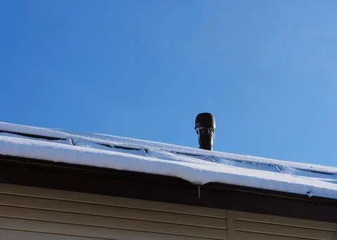 A vent stack with icicles on a snow-covered roof against blue sky background. Foto stock