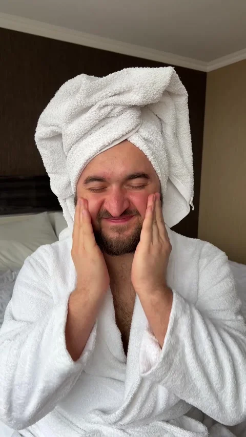 Vertical 4K video. Bearded man in robe, towel on head applies cream to face Stock Footage 268217380