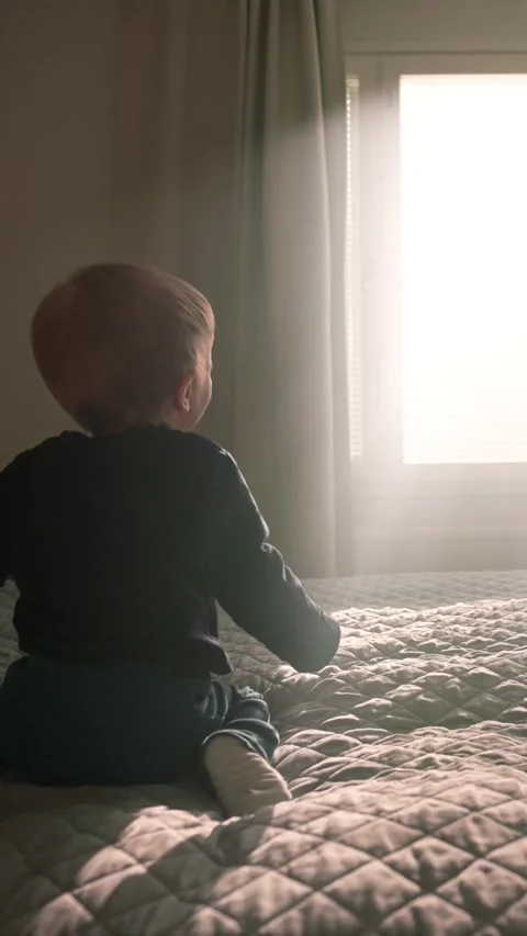 Vertical of Active blond toddler jumps on soft bed looking out of window Stock Footage 274153264