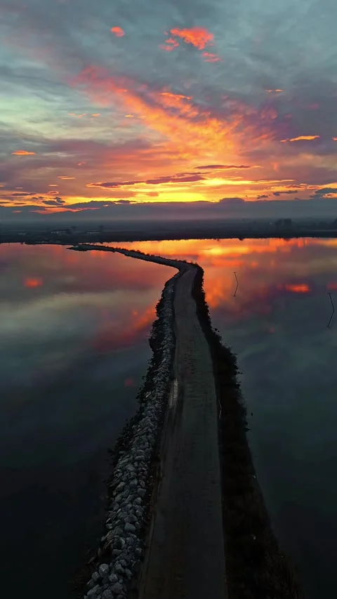 Vertical aerial view of a narrow dirt causeway in a calm lagoon at fiery sunset Stock Footage 345996187