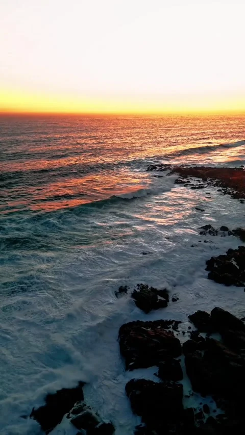 Vertical Aerial View of Ocean Waves Crashing on Rocky Shore at Sunset Stock Footage 337719260
