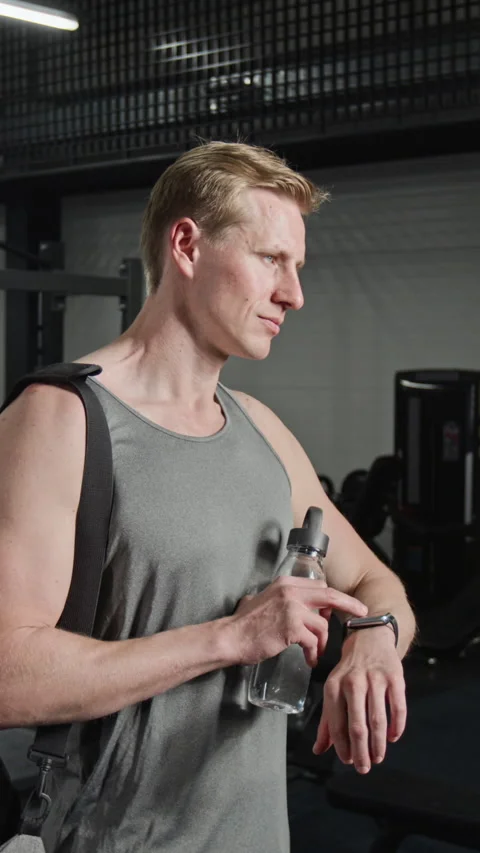 Vertical of Athletic Man Checking Smartwatch with Water Bottle in Gym Stock Footage 339889979