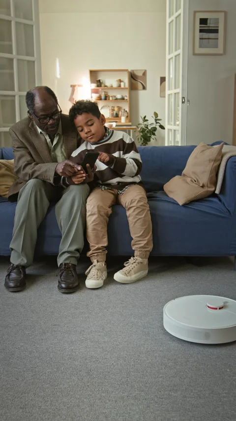 Vertical of Black Child Teaching Granddad to Use Robot Vacuum on Couch Stock Footage 322520141