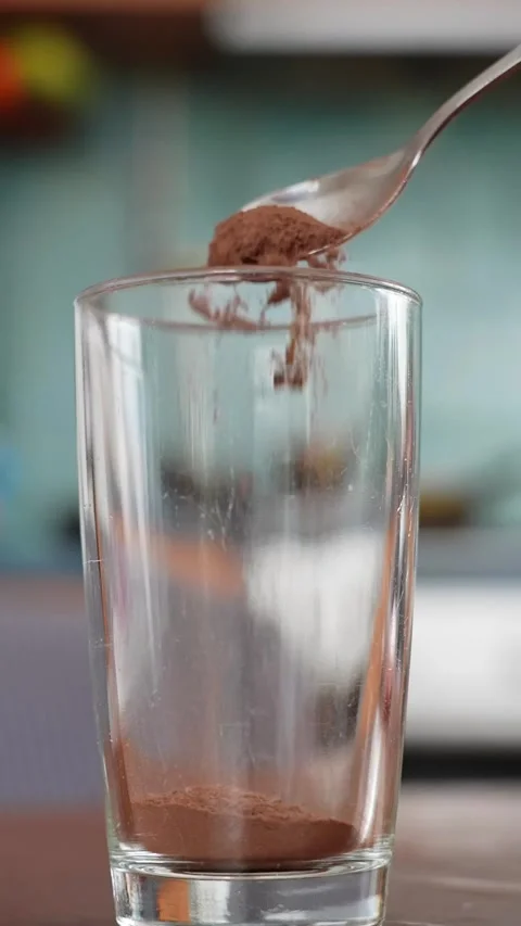 Vertical close-up of cocoa powder falling from a spoon into an empty tall glass Stock Footage 327689874