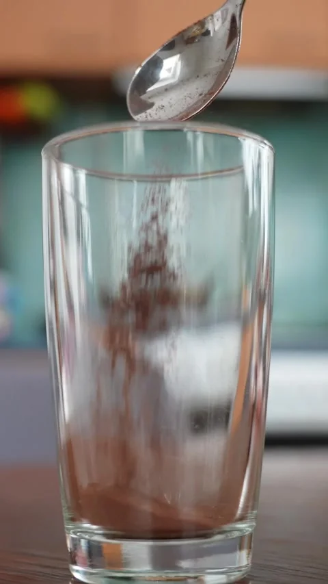Vertical close-up of cocoa powder falling from a spoon into an empty glass on a Stock Footage 328070548