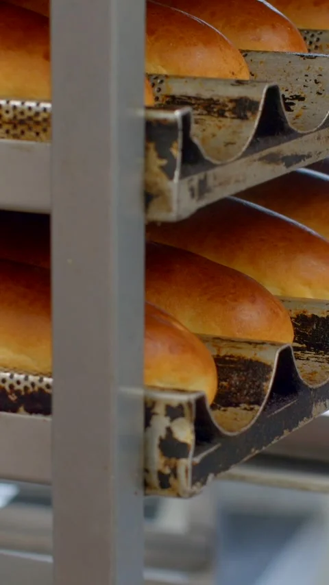 Vertical close-up of stacked golden bread loaves on bakery rack with metal trays Stock Footage 344573642