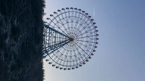 Vertical: dark ferris wheel up the hill on blue sky background. Aircraft trail Stock Footage 233342485
