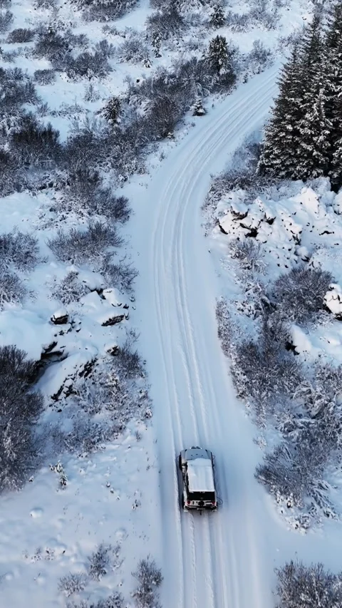 Vertical drone view of a 4x4 driving along a winding snow-covered road through 스톡 동영상 347880154