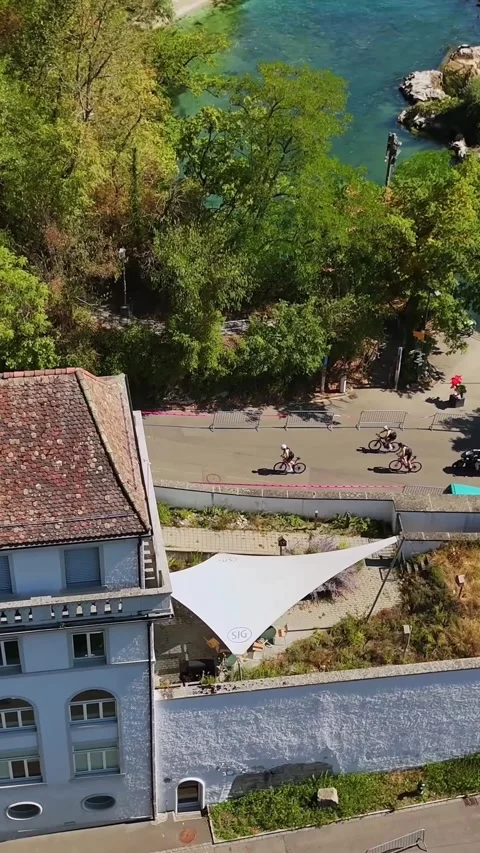 Vertical drone view of cyclists passing a historic riverside building in Vídeos de archivo 346015675