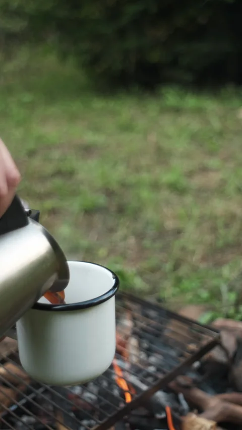 Vertical footage of young man pouring hot coffee sitting near bonfire in nature Stock Footage 285708679