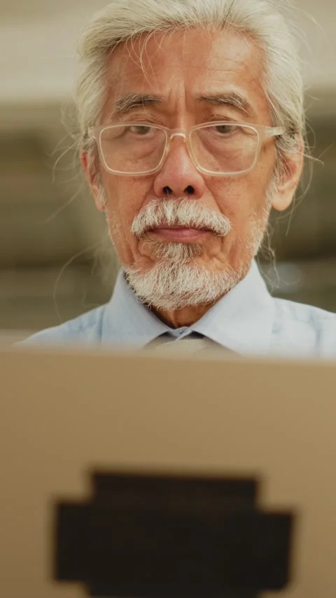 Vertical front close-up of senior Thai man glancing up from laptop screen Video stock 347776785