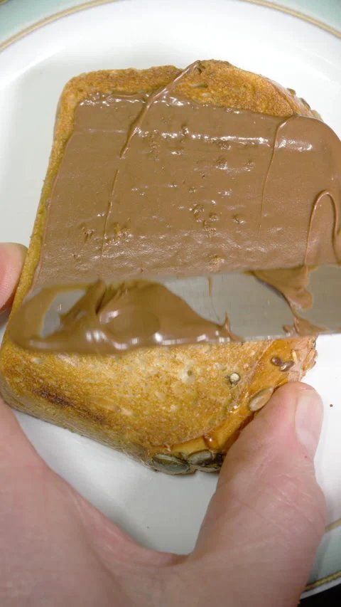 Vertical – A hand with knife spreading a layer of chocolate spread on toast. Video stock 283464449