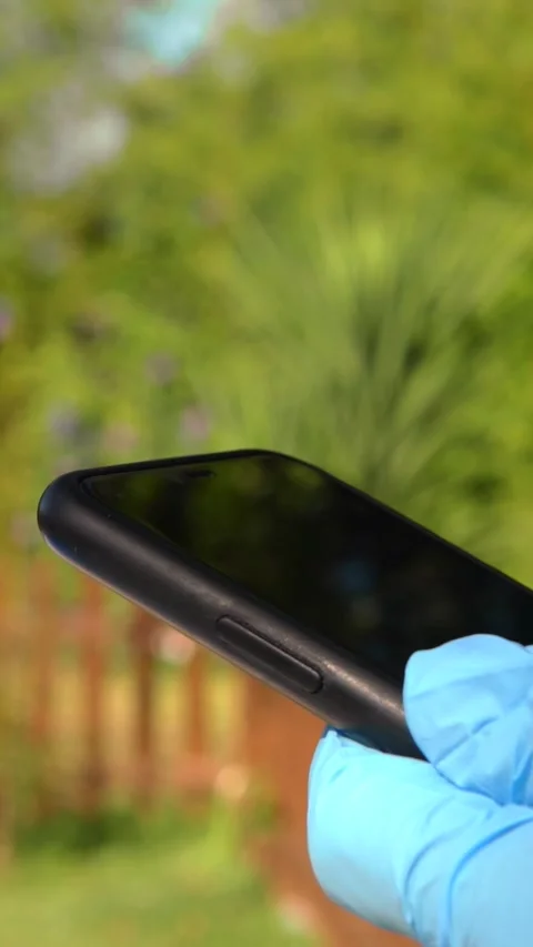 Vertical Hands in blue gloves holding a smartphone in garden. Man using a phone. Stock Footage 314466304
