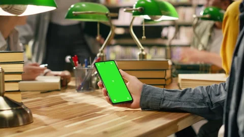 Vertical isolated green screen on smartphone held over wooden library desk Stock Footage 336361727