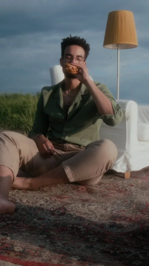 Vertical of Man Eating Hamburger on Beach at Sunset, Sitting on Carpet Stock Footage 304956550