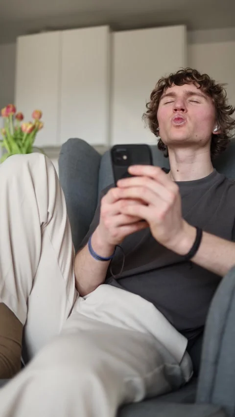 Vertical screen: Bored young man blowing a kiss to his smartphone while sitting  Stock Footage 302632424