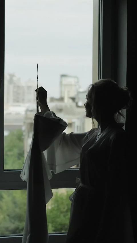 Vertical screen: Bride holding her wedding dress, looking out the window Stock Footage 322051423
