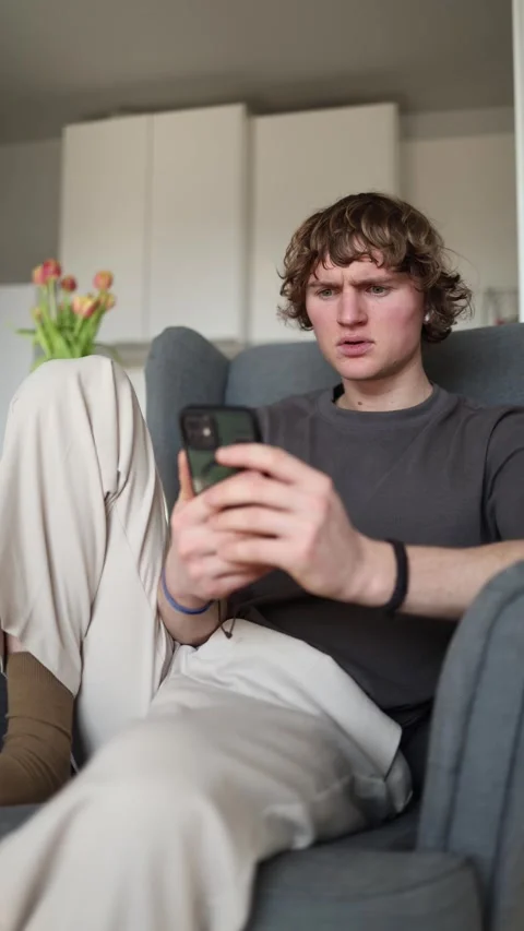 Vertical screen: Confused young man holding smartphone, staring puzzled at the s Stock Footage 302632498