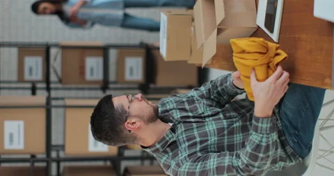 Vertical screen: A male worker boxes a yellow jumper in a room with shelves full Stock Footage 228676669