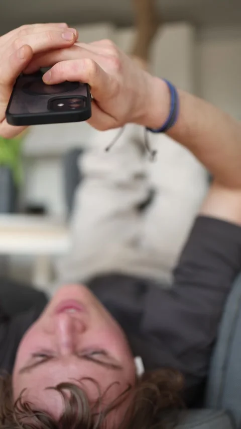 Vertical screen: The man is holding his phone up with both hands while lying dow Stock Footage 302633220