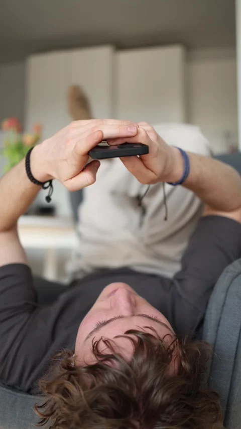 Vertical screen: The man is lying back and playing or browsing something on his  스톡 동영상 302633188