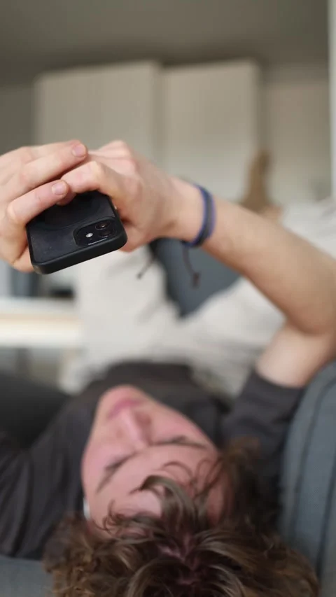 Vertical screen: The man is lying on a couch, absorbed in his phone screen. He h Stock Footage 302633227