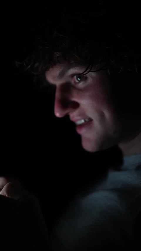 Vertical screen Man smiling while scrolling through phone in the dark his face g Stock Footage 313842824