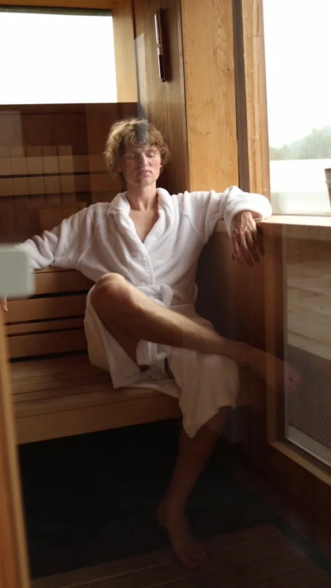 Vertical screen man in white bathrobe sits in wooden sauna by the window enjoyin Stock Footage 318217466