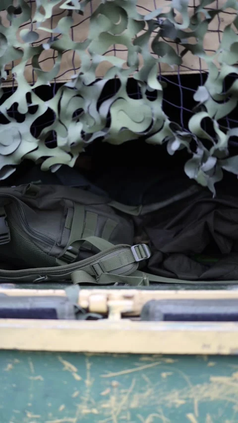 Vertical screen military backpack lies tucked under camouflage netting in rugged Stock Footage 311149915