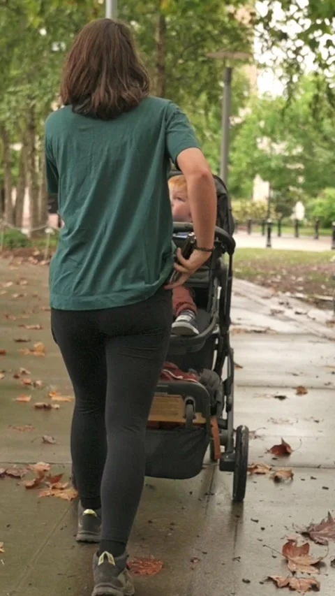 Vertical screen mother adjusting stroller while glancing at phone on leafy park Видео 323575304