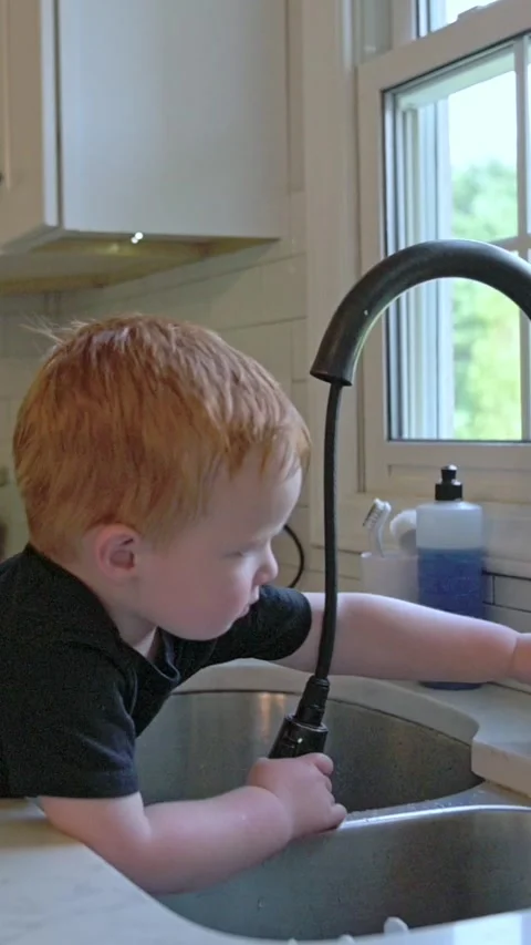 Vertical screen redhaired boy tunes kitchen faucet flow, fairskinned child Stock Footage 323096846
