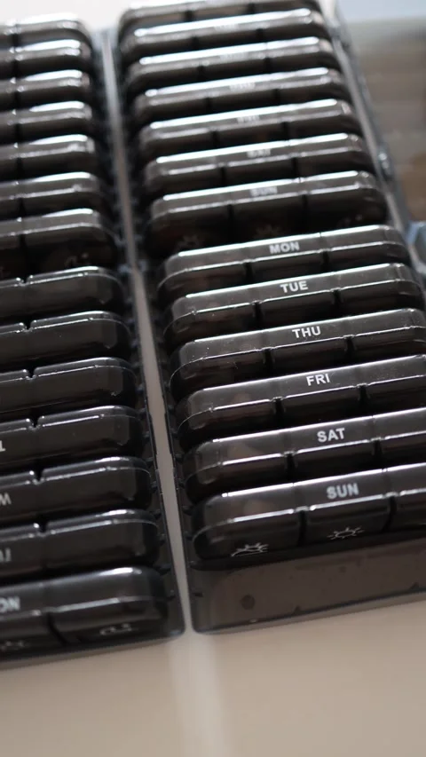 Vertical screen: Rows of black pill organizers labeled with days of the week are Stock Footage 301240194