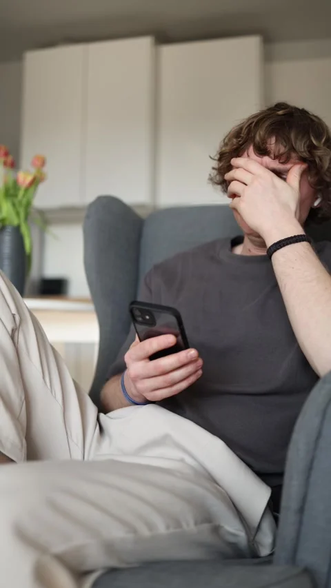 Vertical screen showing a young man sitting in a chair, looking at his smartphon Video stock 302632683