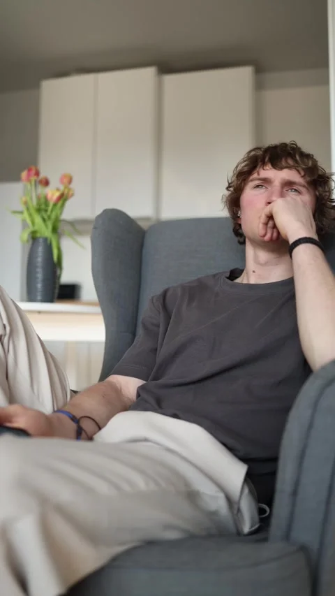 Vertical screen: Thoughtful young man sitting on sofa holding smartphone, deep i Stock Footage 302632369