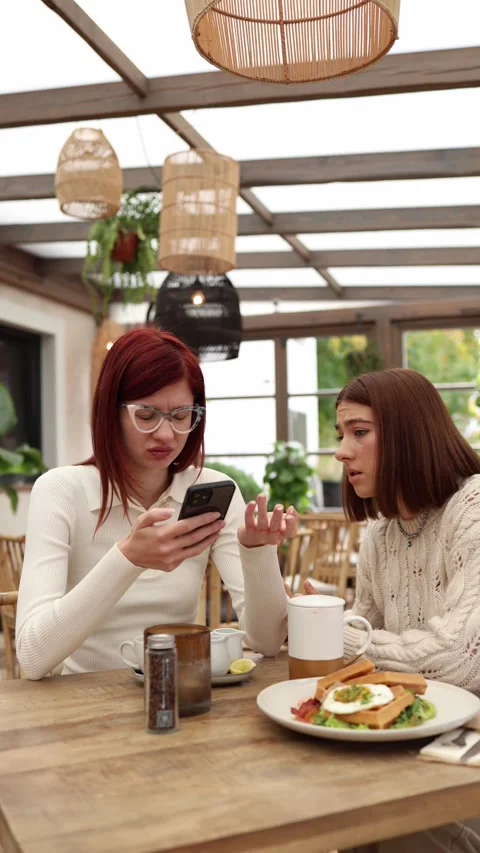 Vertical screen two friends sit in a cozy cafe as one reads a shocking text Stock Footage 320827187
