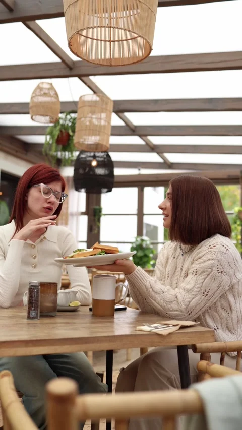 Vertical screen two young women sit in a cafe as one offers her plate  Stock Footage 320827182