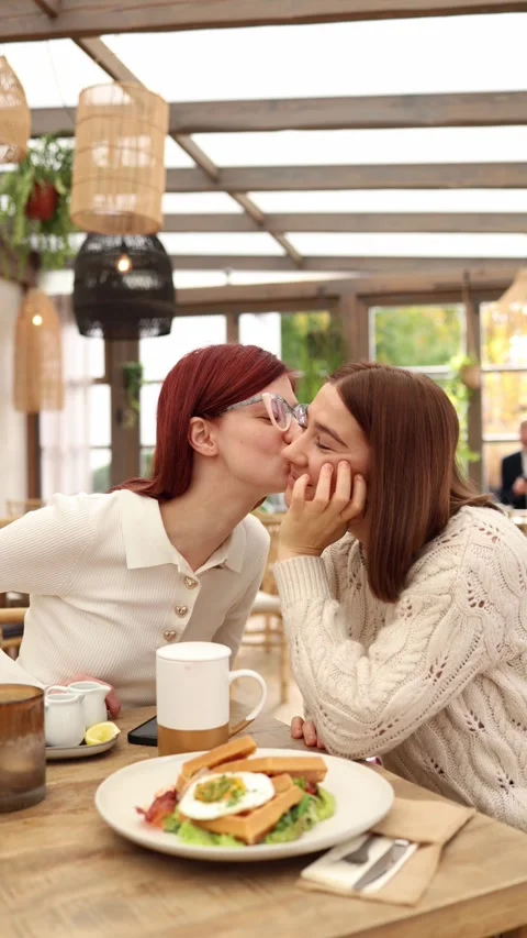 Vertical screen two young women sit in a cozy cafe one leans in to kiss Video stock 320827191