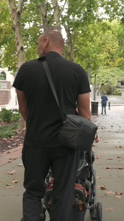 Vertical screen white father pushing stroller on sidewalk under green trees Видео 323575336