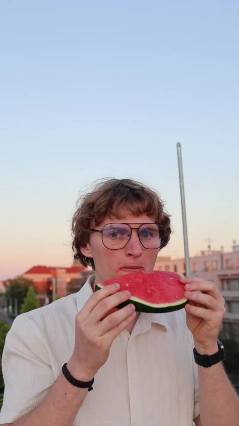 Vertical screen young man in glasses biting fresh watermelon at sunset with eyes Stock Footage 315413734